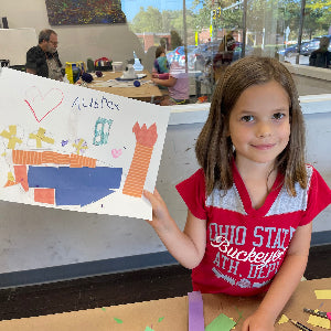 A young girl with brown hair wearing a red and gray 'Ohio State Buckeyes Athlete' t-shirt holds up a colorful handmade paper collage on a table inside a classroom or workshop. Other people and a window are visible in the background.